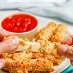 A hand pulling a cheesy breaded mozzarella stick apart in front of a plate of mozzarella sticks with marinara and a text overlay on the photo