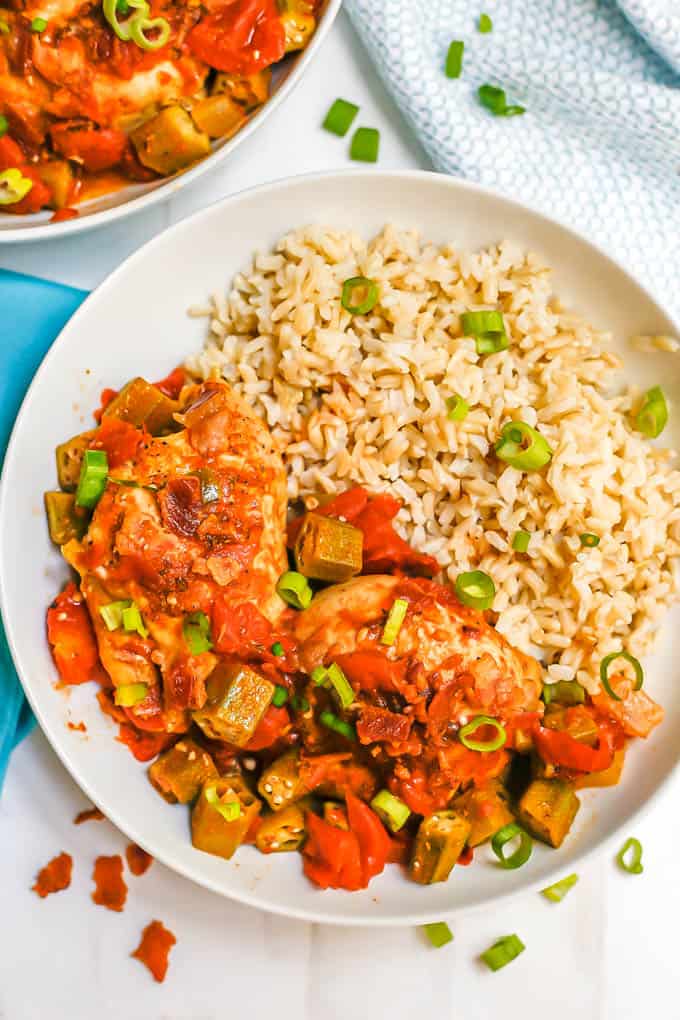 A white bowl with brown rice served alongside Cajun chicken thighs with okra and tomatoes and sliced green onions scattered on top