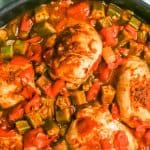 Close up of seared seasoned Cajun chicken and okra in a skillet with tomatoes and veggies with a text overlay on the photo