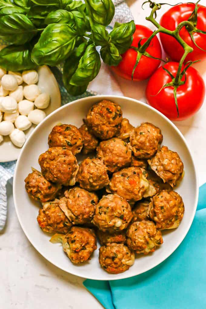 Cheesy meatballs piled in a low white bowl with fresh mozzarella, basil and tomatoes in the background