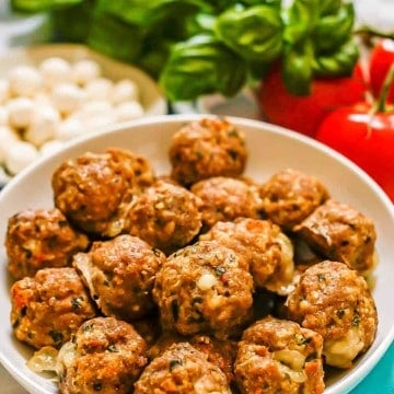 A large low white bowl with cheesy meatballs and fresh mozzarella, basil and tomatoes in the background