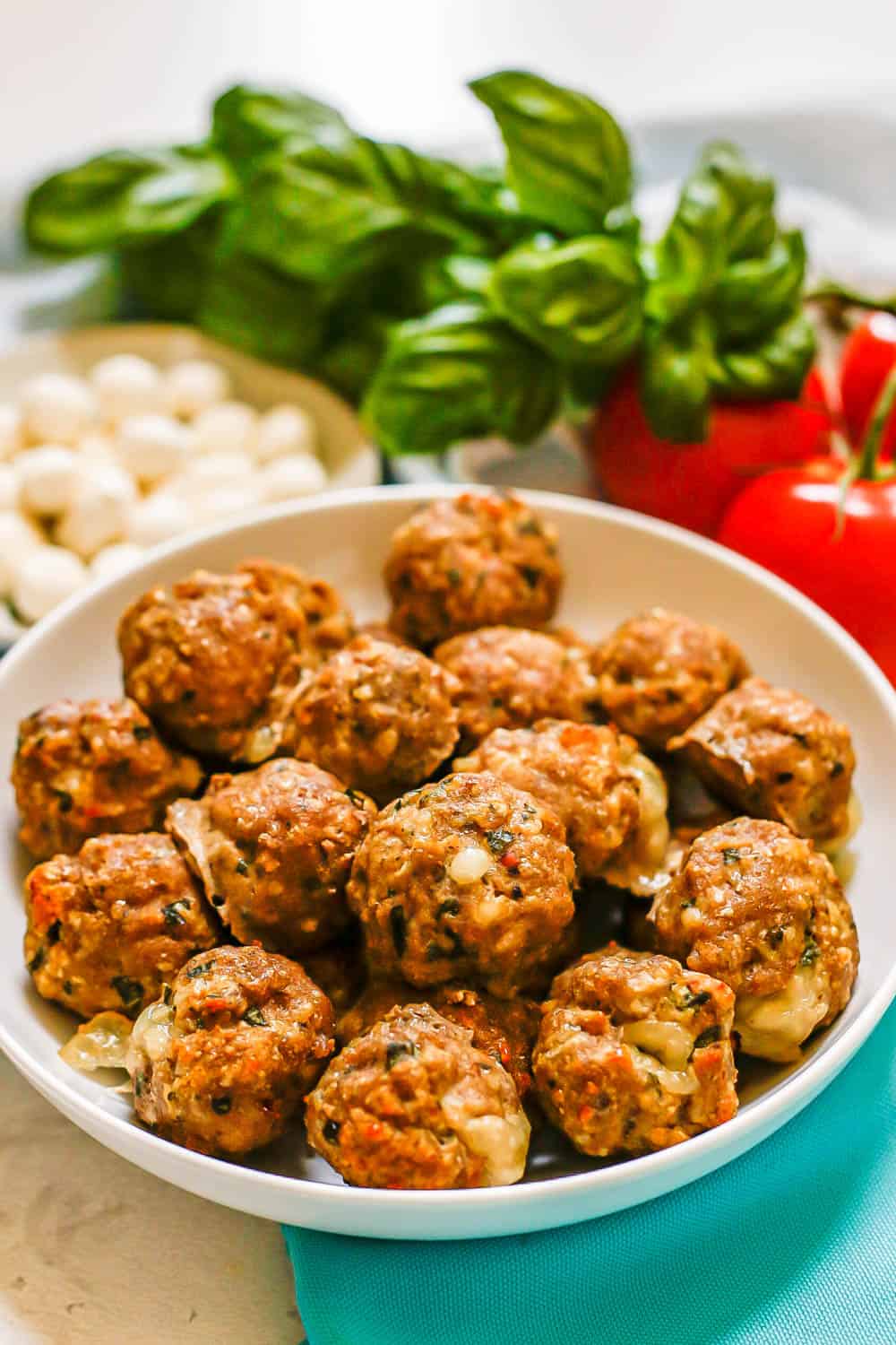 A large low white bowl with cheesy meatballs and fresh mozzarella, basil and tomatoes in the background