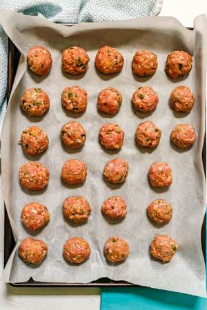 Rolled meatballs lined up on a parchment paper lined baking sheet before going in the oven