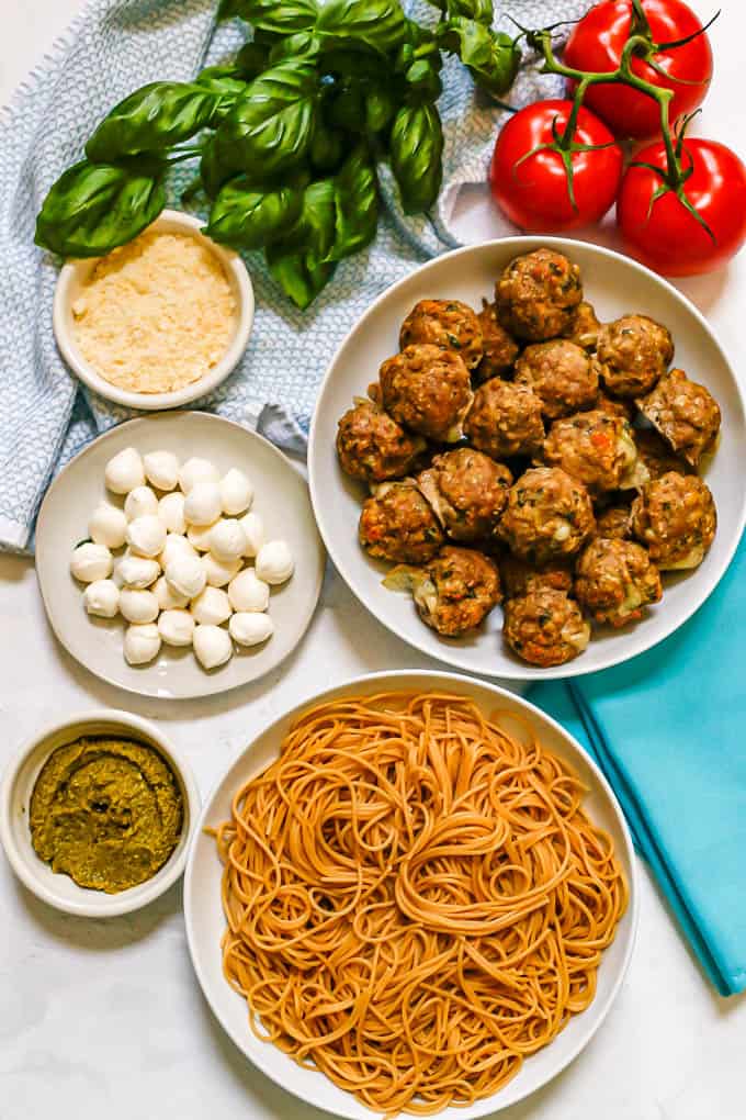 A Caprese meatballs spread with a bowl of meatballs, a bowl of cooked spaghetti noodles, mini mozzarella balls, Parmesan cheese, a bowl of pesto, fresh basil and vine ripe tomatoes