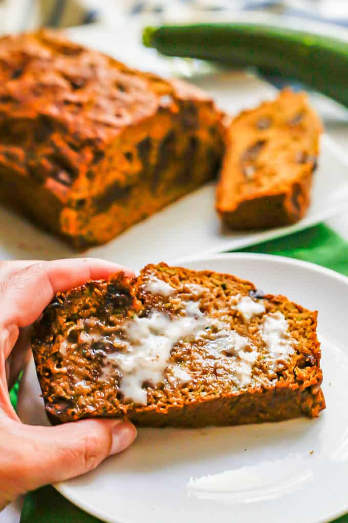 A hand holding up a piece of buttered zucchini bread from a white circular plate with the rest of the loaf in the background