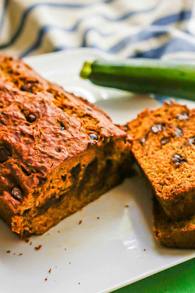A loaf of zucchini bread with chocolate chips on a white plate with a couple of slices stacked to the side
