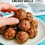 Chocolate peanut butter energy balls piled in a pyramid on a small white plate with a hand reaching for the top one and a text overlay on the photo.