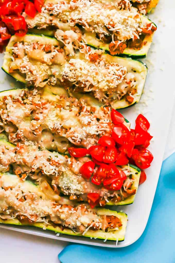 Close up of cheesy sausage stuffed zucchini arranged on a rectangular white platter with piles of fresh tomato on top for serving