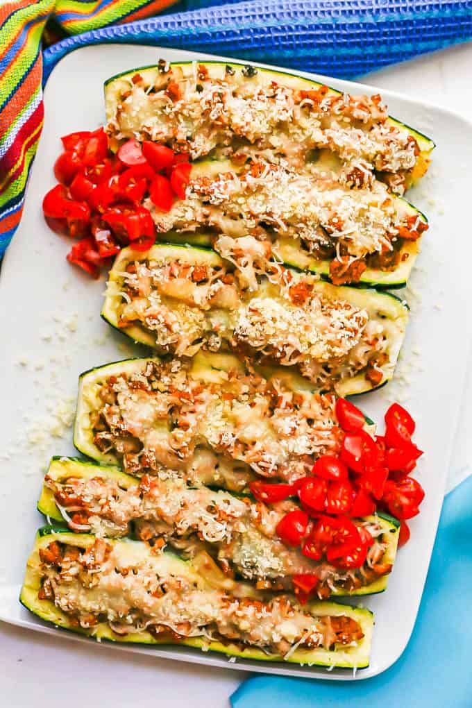 Cheesy sausage stuffed zucchini arranged on a rectangular white platter with piles of fresh tomato on top for serving