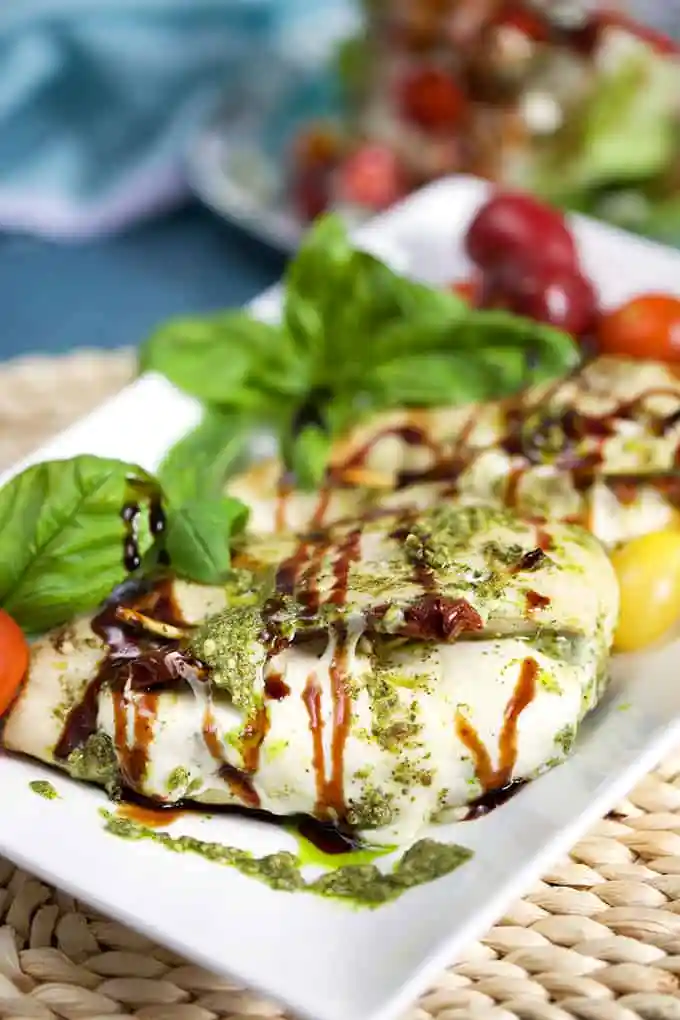 A white platter with Caprese stuffed grilled chicken breasts with pesto and balsamic drizzled on top