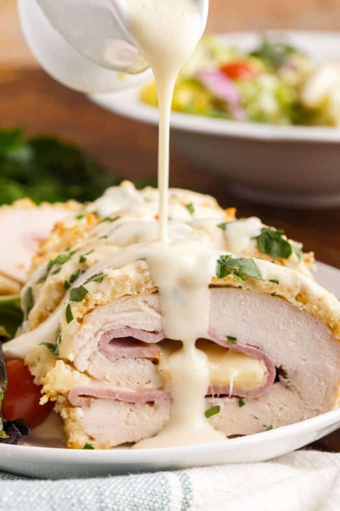 A creamy sauce being poured over chicken cordon blue on a white plate