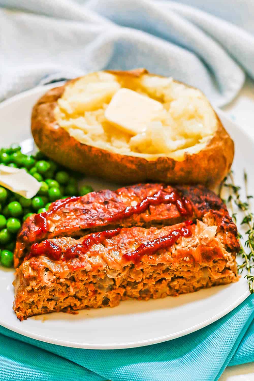Ground turkey meatloaf slices served on a white plate with a baked potato and green peas