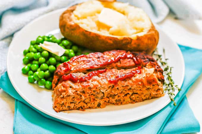 A white dinner plate with a baked potato, green peas and two slices of turkey meatloaf