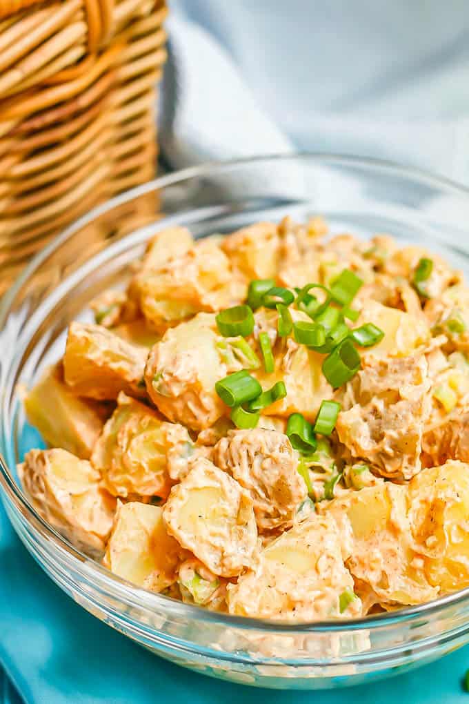 Close up of potato salad in a glass bowl with green onions on top and a picnic basket in the background