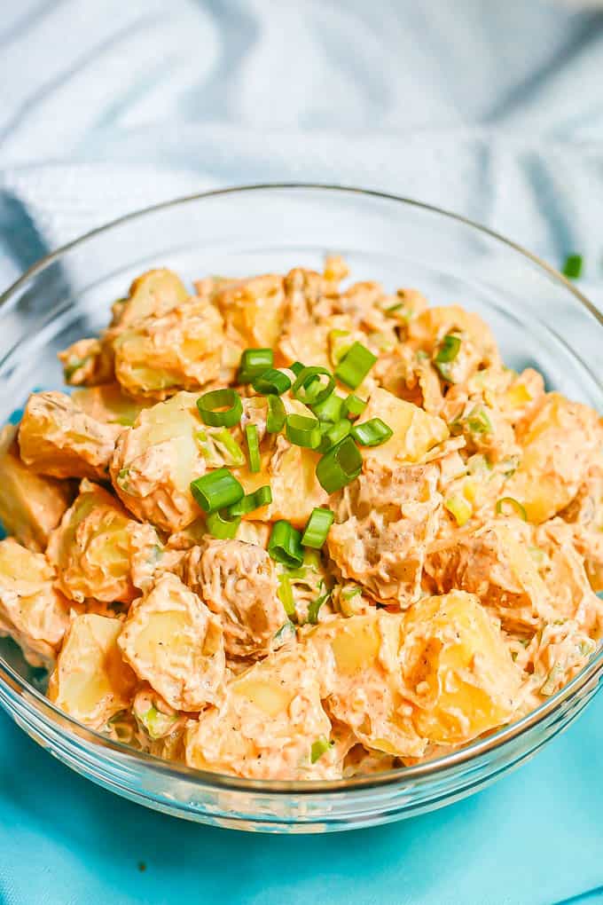 A healthy creamy potato salad in a glass bowl with green onions on top and turquoise napkins to the side