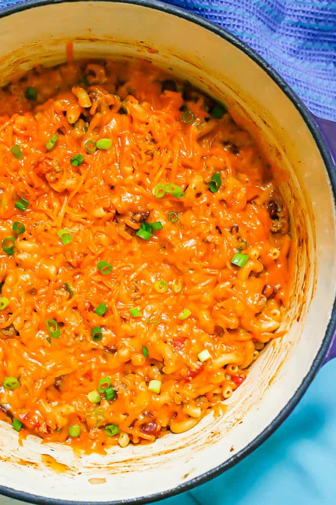 Close up of ground turkey chili mac in a large Dutch oven with a layer of melted cheddar cheese on top and a sprinkling of sliced green onions