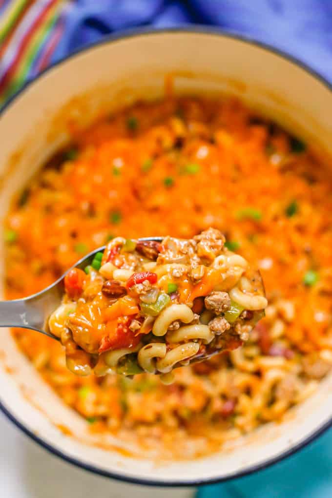 A silver serving spoon lifting a cheesy scoop of turkey chili mac from a large Dutch oven