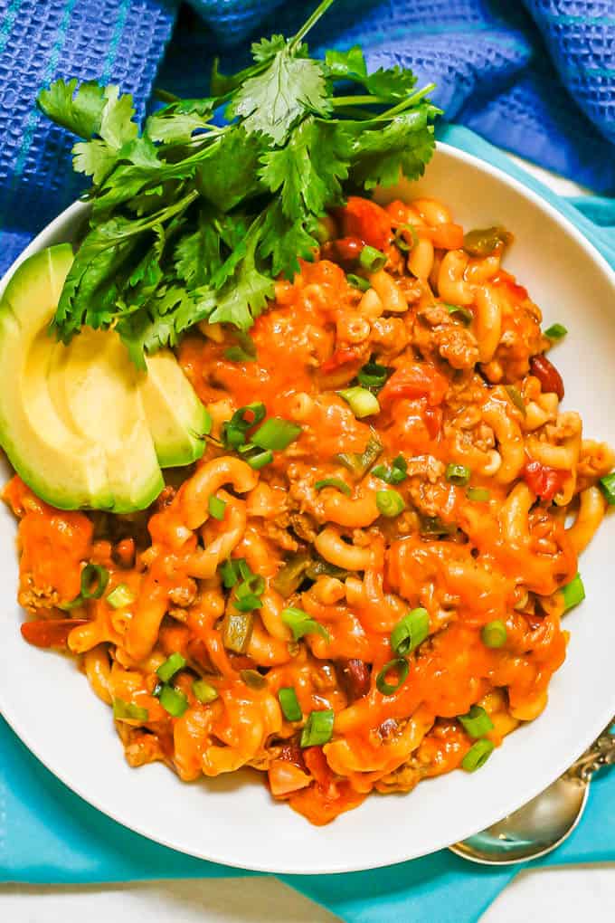 A white bowl with a serving of cheesy chili mac with avocado, cilantro and sliced green onions for toppings