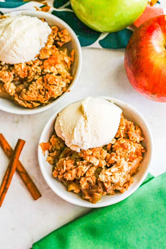 Two small white bowls with apple crisp topped with vanilla ice cream with apples and cinnamon sticks to the side