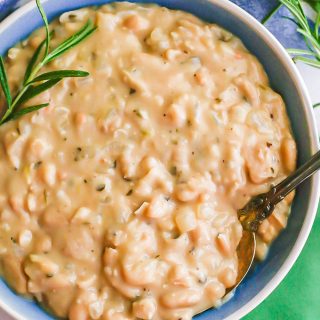 A spoon resting in a blue and white bowl of creamy white beans with rosemary sprigs to the side