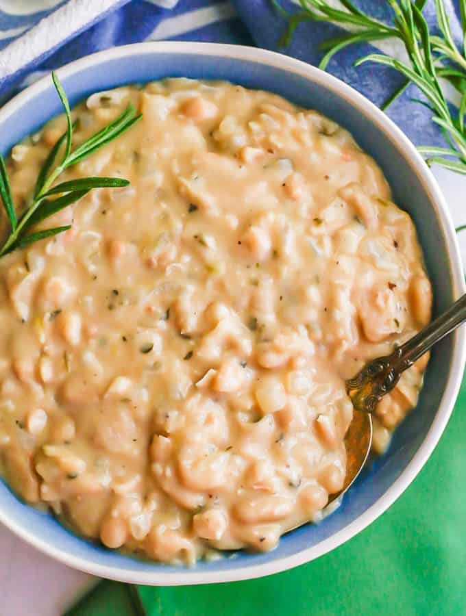 A spoon resting in a blue and white bowl of creamy white beans with rosemary sprigs to the side