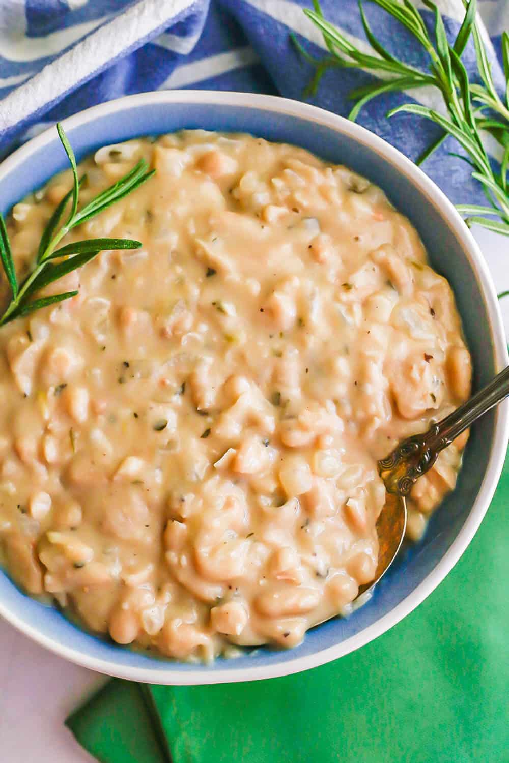 A spoon resting in a blue and white bowl of creamy white beans with rosemary sprigs to the side