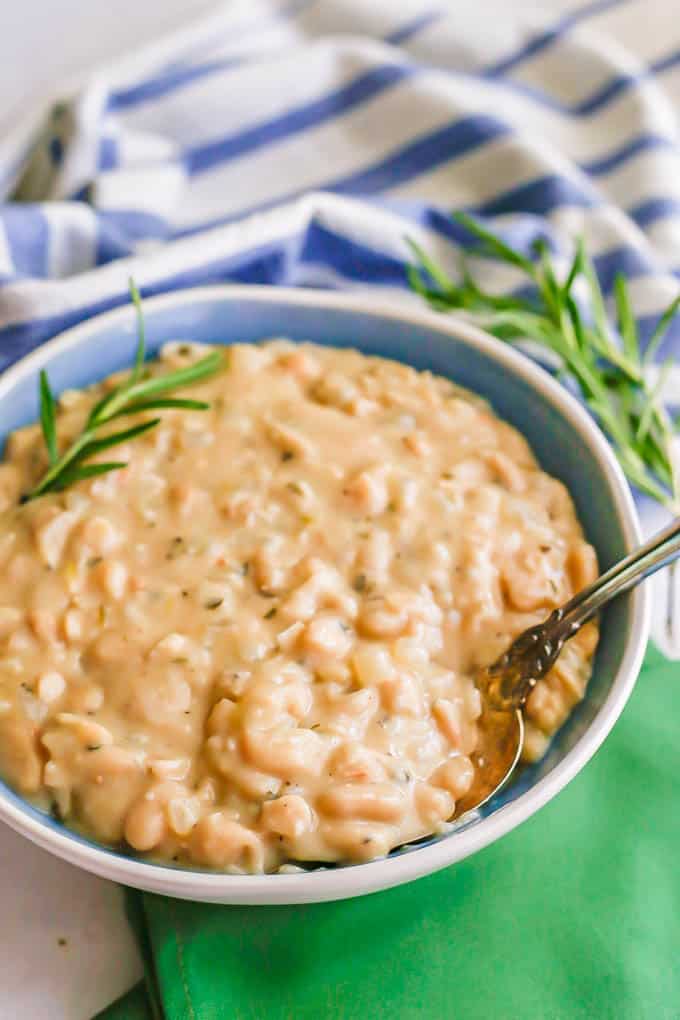 A spoon resting in a blue and white serving bowl of creamy cannellini beans with sprigs of fresh rosemary to the side