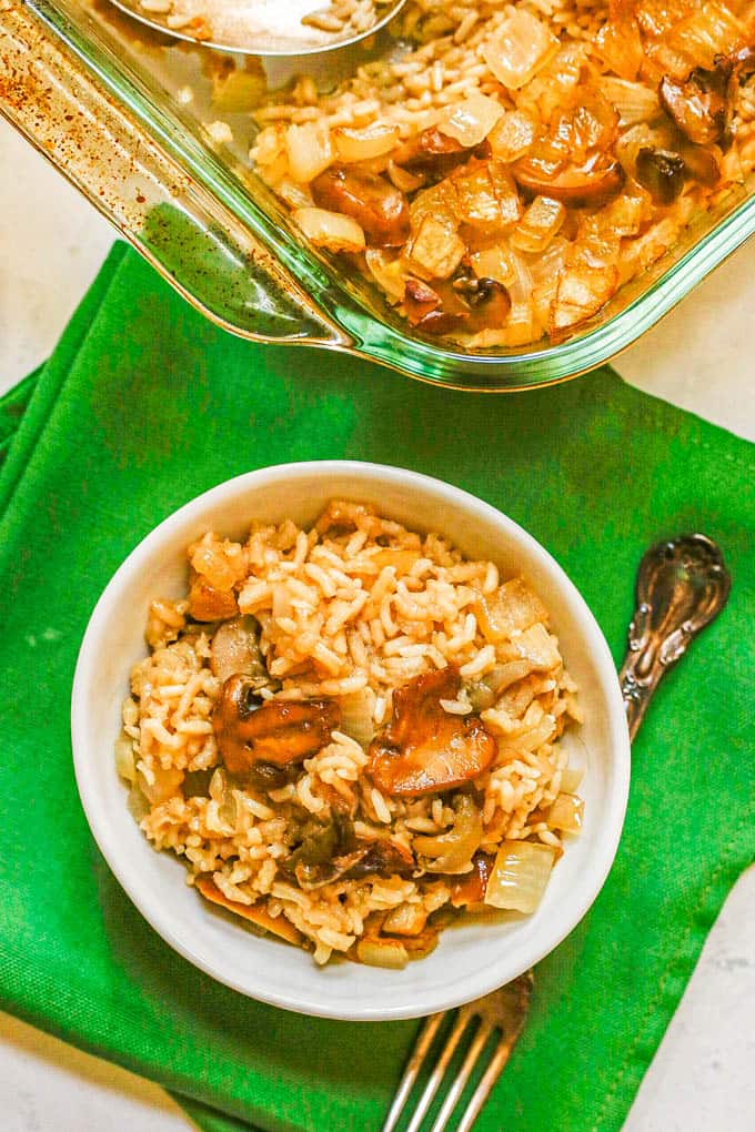 A serving of mushroom rice casserole in a small white dish set on green napkins in front of the glass casserole pan