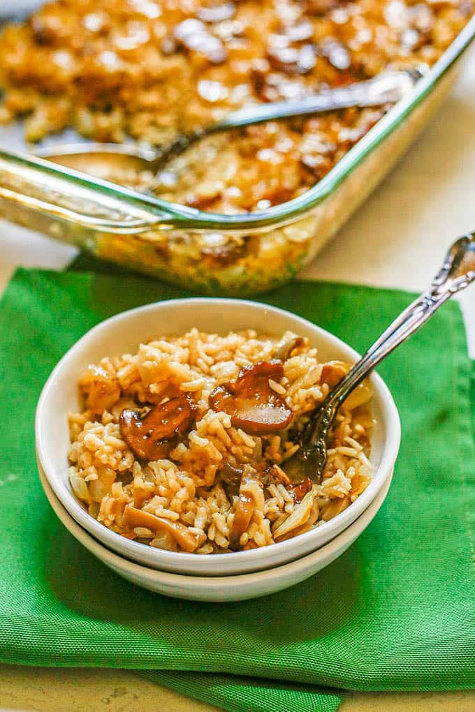 A serving of mushroom rice casserole in a small white dish with a fork tucked in set on green napkins in front of the glass casserole pan