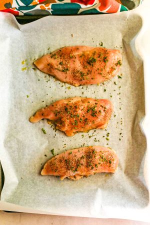 A parchment paper lined baking sheet with 3 chicken breasts seasoned with salt, pepper and fresh rosemary before being roasted