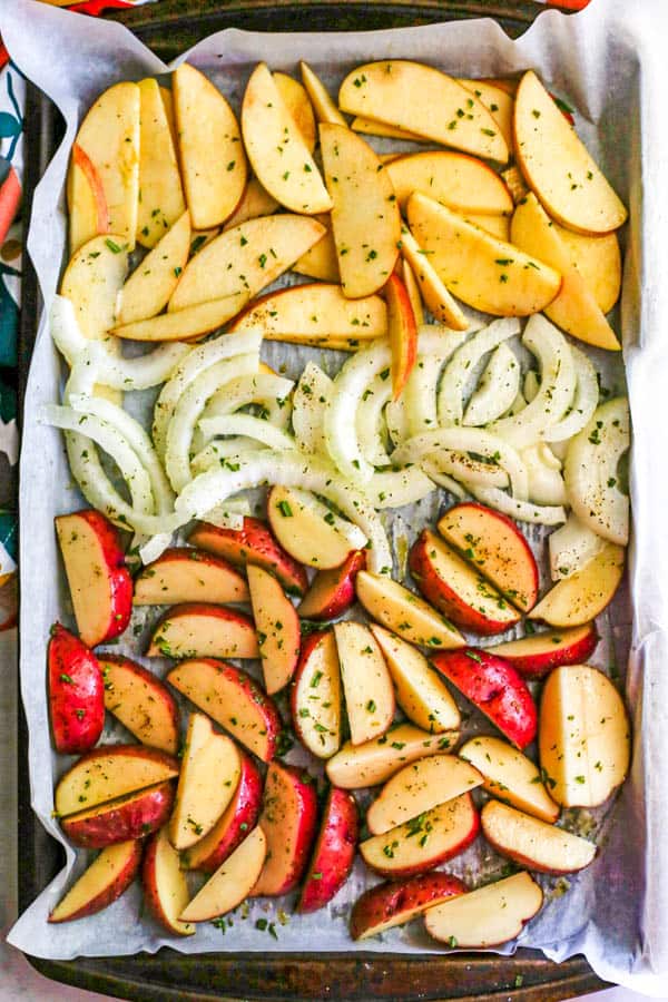 A parchment paper lined baking sheet with rows of sliced apples, onions and red potatoes before being roasted