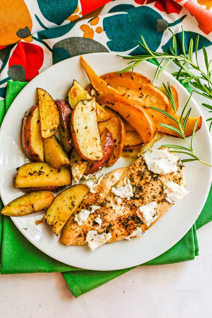 Overhead shot of a round white dinner plate with roasted potatoes, apples and a chicken breast topped with goat cheese with sprigs of rosemary to the side
