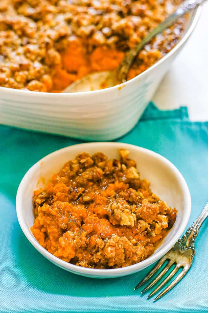 A small bowl with a serving of sweet potato casserole set on teal napkins with a fork nearby and the casserole dish in the background