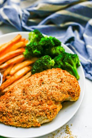 Close up of a white dinner plate with baked Ranch chicken, steamed broccoli and oven fries with a blue striped kitchen towel in the background