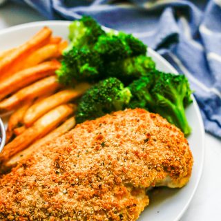 Close up of a white dinner plate with baked Ranch chicken, steamed broccoli and oven fries with a blue striped kitchen towel in the background