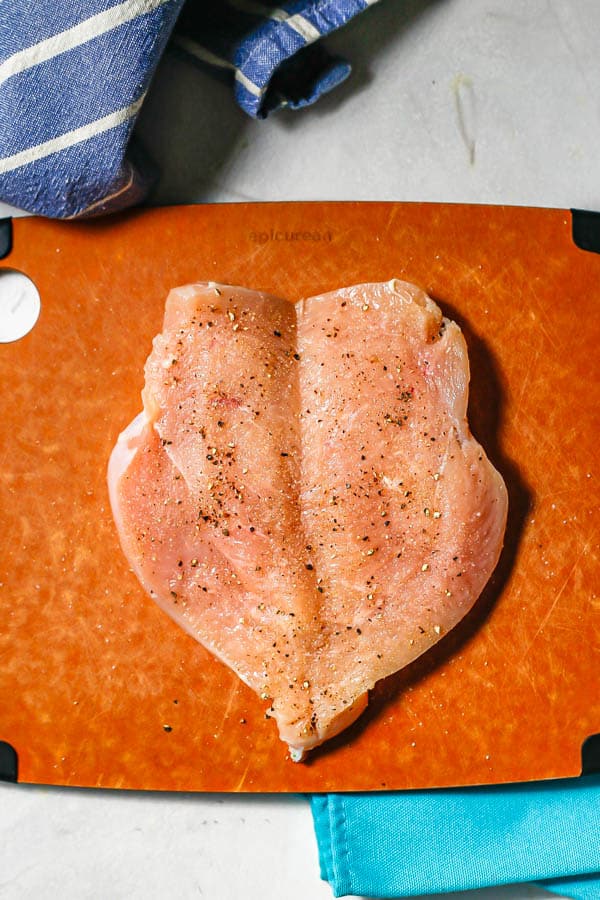 A butterflied chicken breast laying open on a cutting board and sprinkled with salt and pepper.