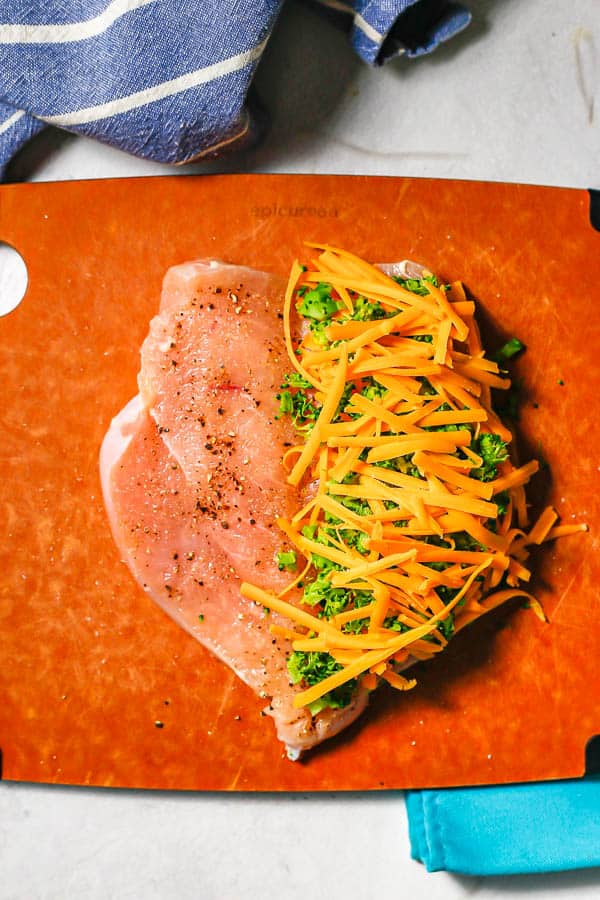 A butterflied chicken breast laying open on a cutting board with shredded cheddar cheese and chopped broccoli piled on one side.