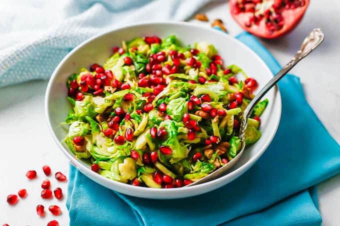 A white bowl with a mix of Brussels sprouts, pecan and pomegranate arils with a serving spoon resting in the bowl