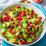 A festive mix of holiday Brussels sprouts with toasted pecans and pomegranate arils in a white serving bowl set on turquoise napkins with a text overlay on the image