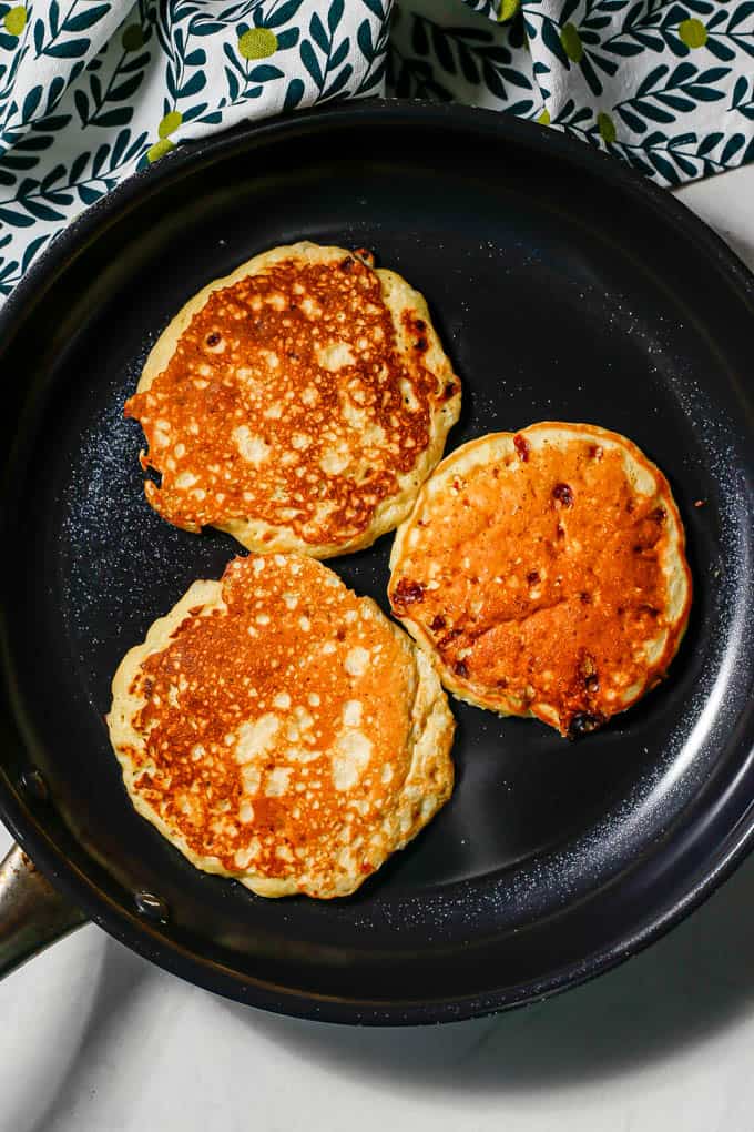 Three pancakes cooking in a dark nonstick skillet.