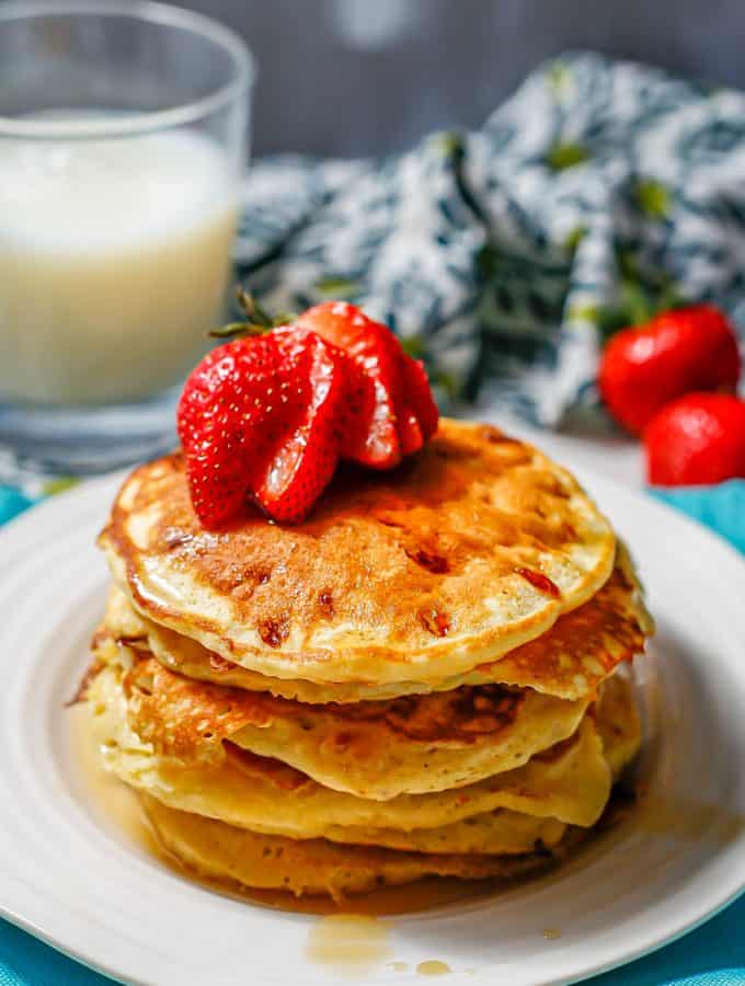 Cottage cheese pancakes stacked on a white plate with maple syrup drizzled over and a sliced strawberry on top.