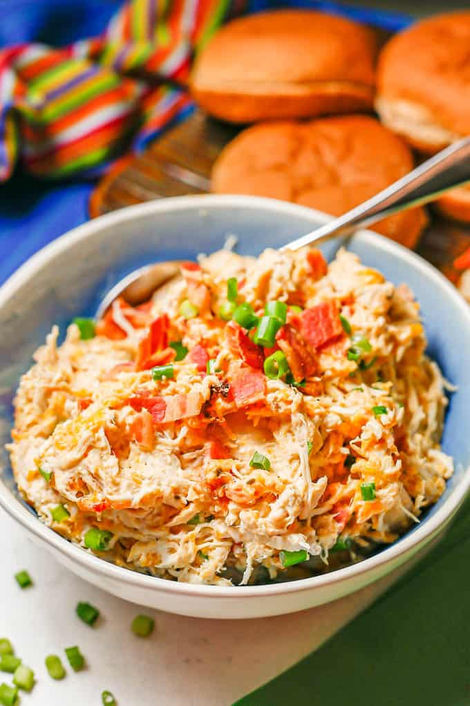 A spoon resting in a bowl of a creamy, cheesy shredded chicken mixture with green onions and bacon on top.