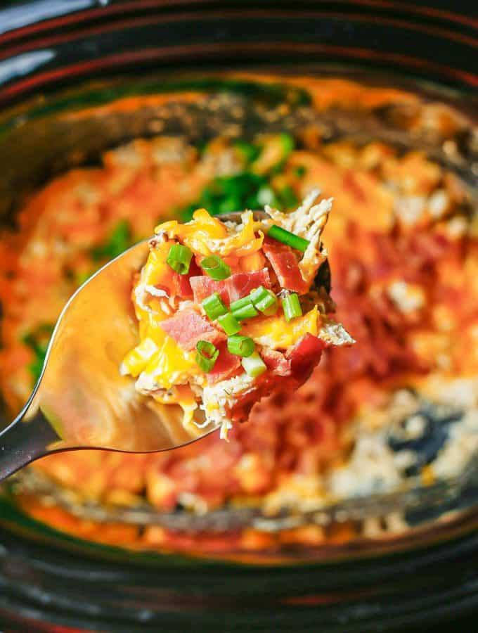 A silver spoon lifting up a scoop of crock pot crack chicken with bacon and green onions from the slow cooker insert.