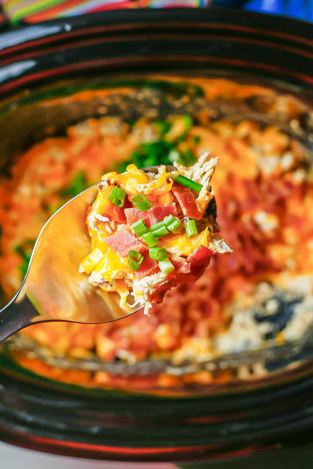 A silver spoon lifting up a scoop of crock pot crack chicken with bacon and green onions from the slow cooker insert.