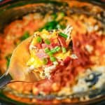 A silver spoon lifting up a scoop of crock pot crack chicken with bacon and green onions from the slow cooker insert with a text overlay on the photo.