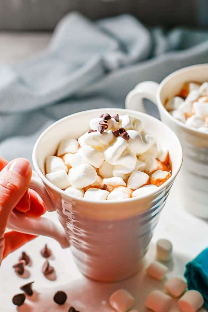 A hand picking up a coffee mug full of hot cocoa topped with mini marshmallows, whipped cream and mini chocolate chips