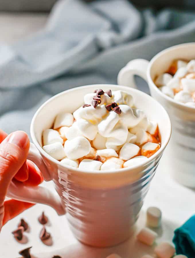 A hand picking up a coffee mug full of hot cocoa topped with mini marshmallows, whipped cream and mini chocolate chips