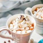 Hot chocolate served in white mugs and topped with mini marshmallows, whipped cream and mini chocolate chips with a text overlay on the photo