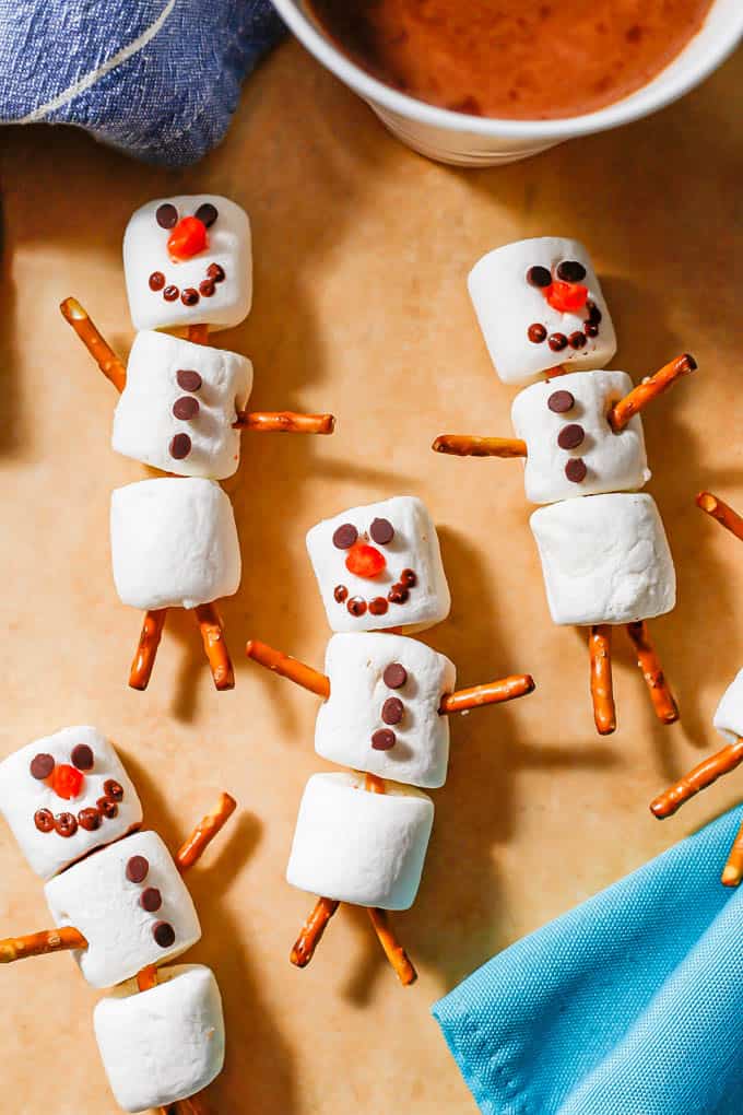 Decorated marshmallow snowmen on parchment paper near a mug of hot cocoa