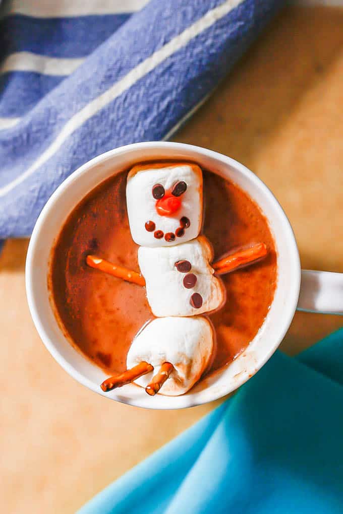A marshmallow snowman with pretzels for arms and legs floating in a mug of hot chocolate